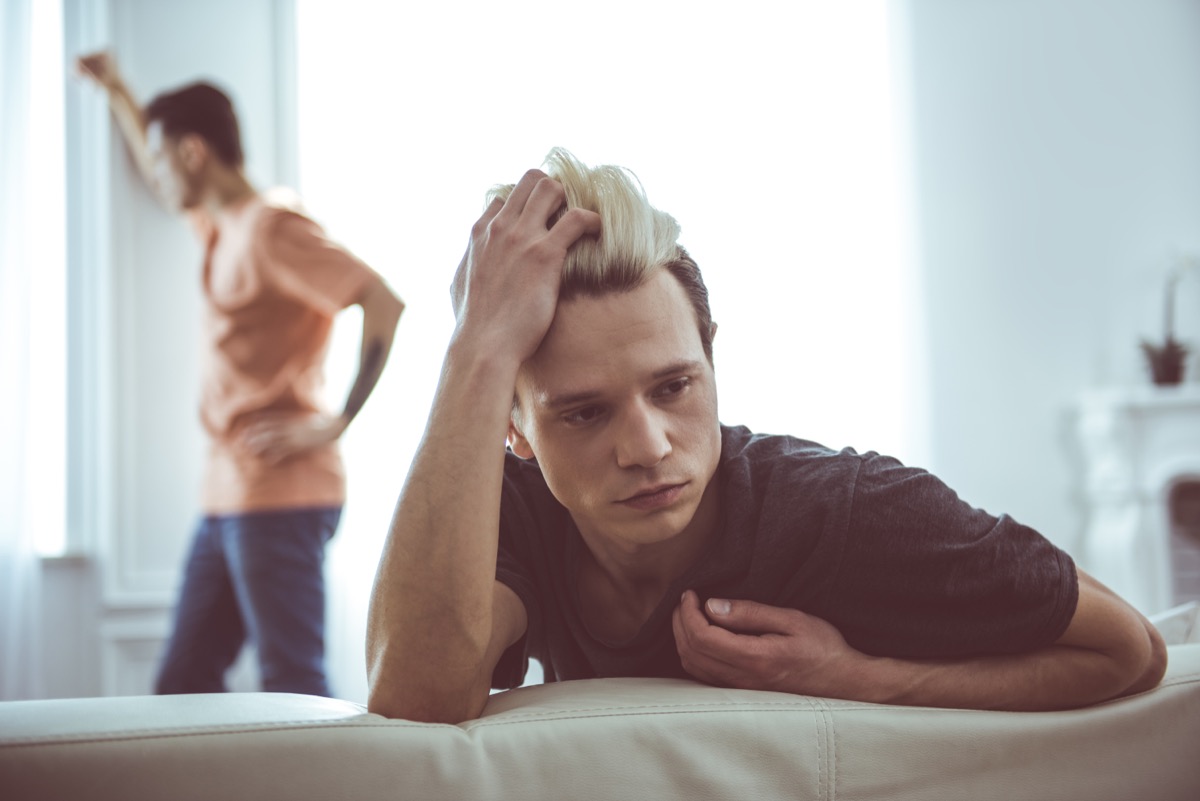 Toned portrait of sad guy with dyed hair sitting on couch while his boyfriend standing near window on blurred background