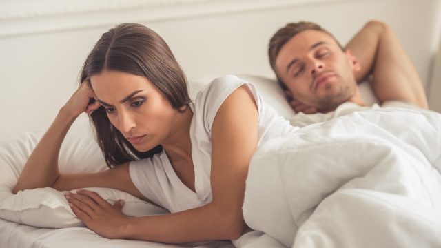 Offended woman is leaning on her hand and looking down while her husband is sleeping. Both are lying in bed