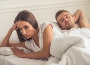 Offended woman is leaning on her hand and looking down while her husband is sleeping. Both are lying in bed