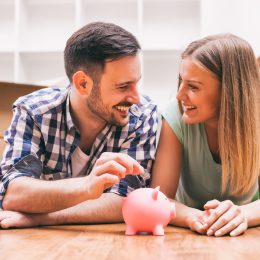 A couple lying on the floor in a home surrounded by moving boxes while putting money into a piggy bank