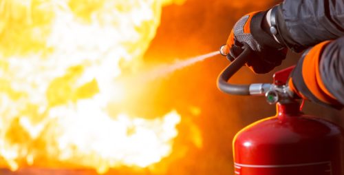 A close-up showing a person pointing a fire extinguisher nozzle at the base of a large fire.