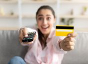 Closeup of excited millennial woman sitting on couch in living room, using tv remote and holding yellow credit card