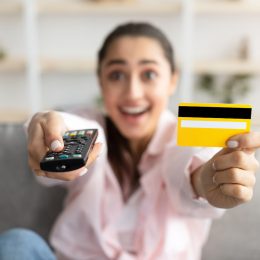 Closeup of excited millennial woman sitting on couch in living room, using tv remote and holding yellow credit card