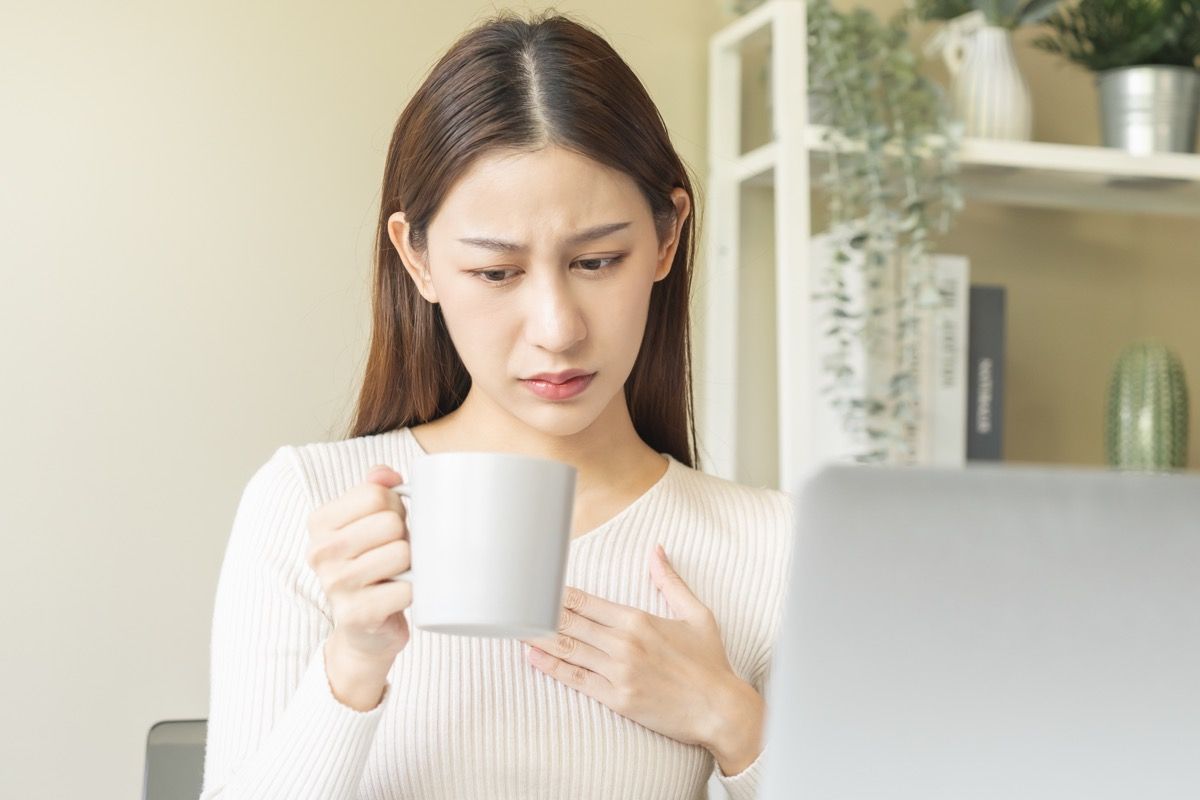 Palpitation, suffer asian young woman holding mug, drinking strong a cup of coffee, touching her chest after drink caffeine, tea while working from home, face expression stress, illness people.