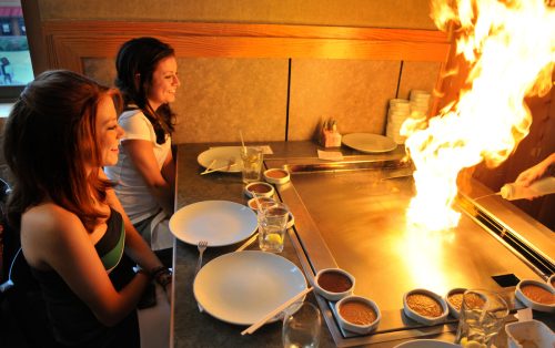 Two young girls sitting at a Hibachi restaurant with the grill flaming up.