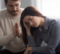 Husband sitting on couch with his wife and pleading for forgiveness with hands in prayer.