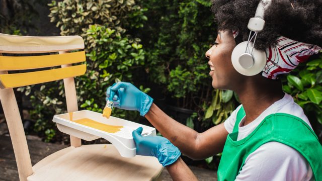 A young woman is painting vintage chair in yellow color with a paintbrush in the back yard while listening to music via wireless headphones