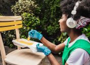 A young woman is painting vintage chair in yellow color with a paintbrush in the back yard while listening to music via wireless headphones