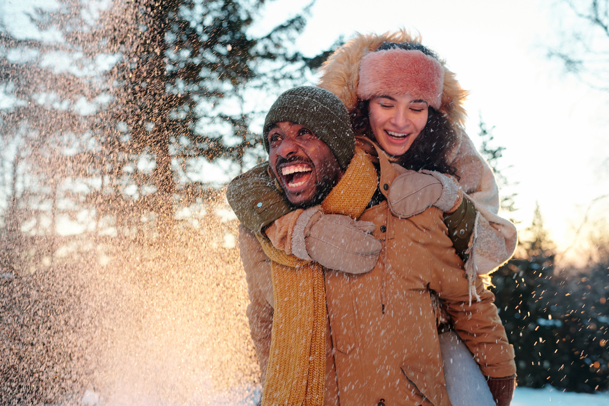 Cheerful multiracial couple in winterwear laughing while girl embracing her boyfriend during snowfall