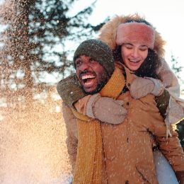 Cheerful multiracial couple in winterwear laughing while girl embracing her boyfriend during snowfall