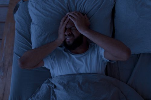 Sleeping Problem. Overhead above top view of stressed African American guy lying alone in bed touching grabbing head feeling depressed, suffering from insomnia