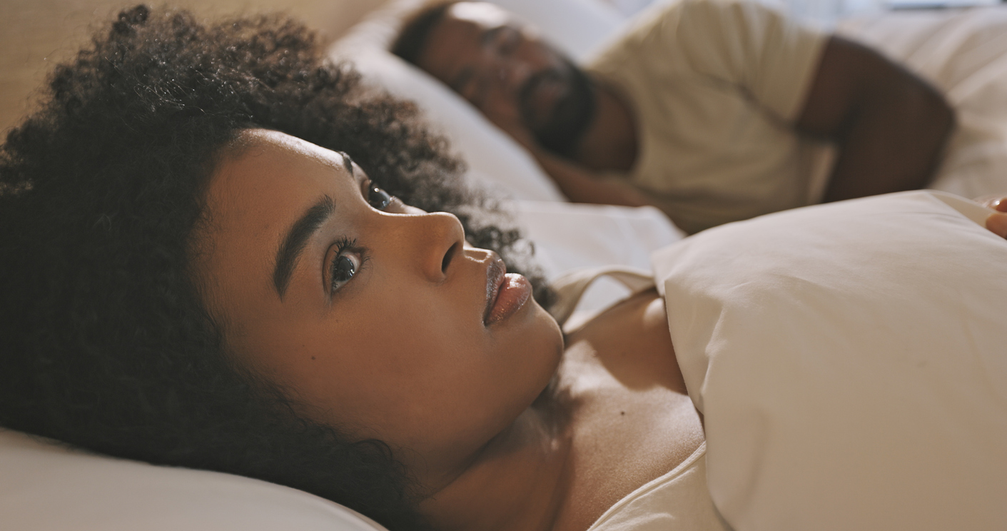 Woman lying in bed with her partner, waking up and looking anxious. 