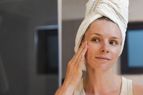 Woman with her hair wrapped in a towel applying eye cream in the mirror.