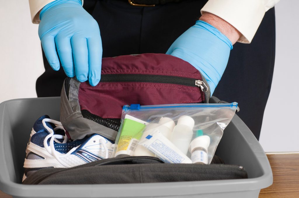 Gloved hands of an airport security person examining the contents of a bin with a traveler's belongings.
