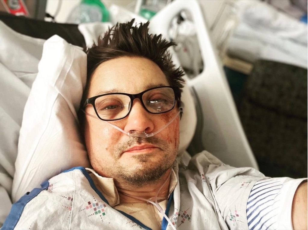 Jeremy Renner sitting in a hospital bed wearing glasses and an oxygen tube