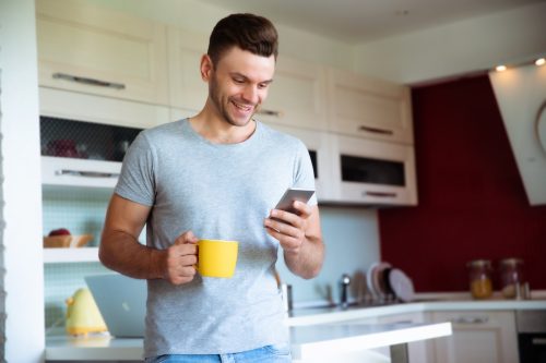 A young man in a gray t-shirt standing in the kitchen with a yellow coffee mug in one hand and his cell phone in the other