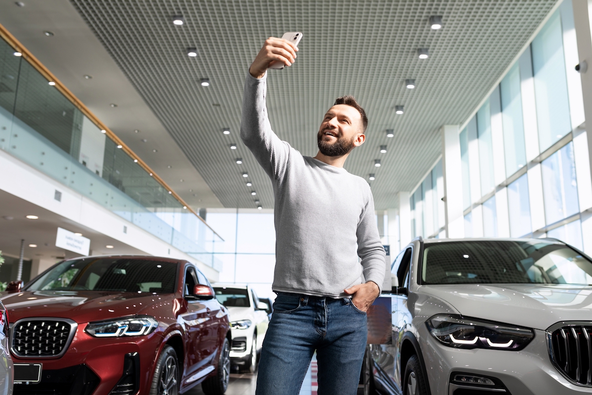 A middle-aged man taking a selfie in a car dealership to show off.