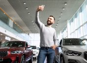 A middle-aged man taking a selfie in a car dealership to show off.