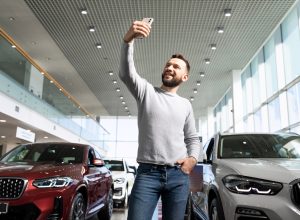 A middle-aged man taking a selfie in a car dealership to show off.