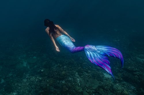 Young female free diver swims underwater in a colorful mermaid costume.