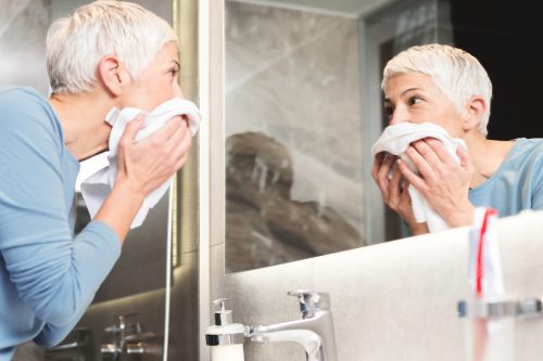 Mujer mayor mirándose en el espejo y secándose la cara con una toalla.