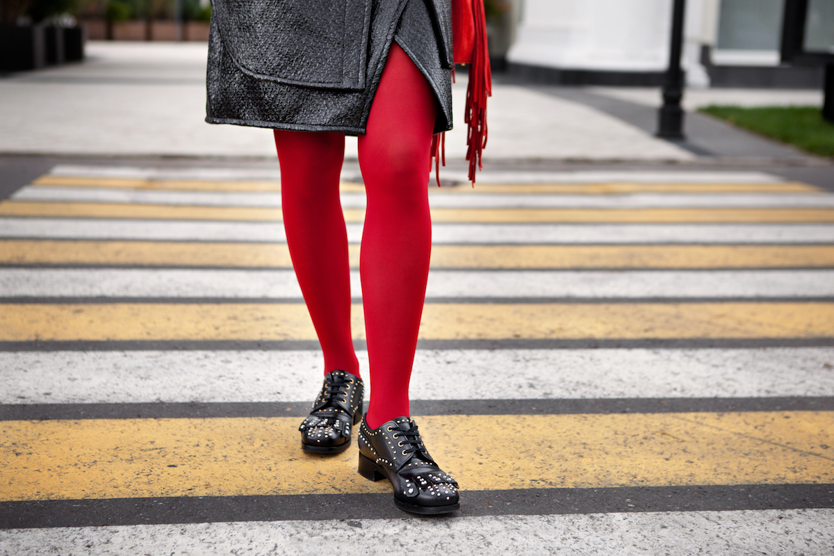 Woman legs in red color tights, black leather shoes with rivets and skirt stand on crosswalk. Fashion details for stylish extravagant women