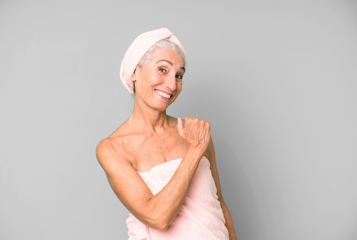Older woman with gray hair wrapped in a light pink towel and matching hair towel after a shower, smiling and looking happy.