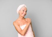 Older woman with gray hair wrapped in a light pink towel and matching hair towel after a shower, smiling and looking happy.