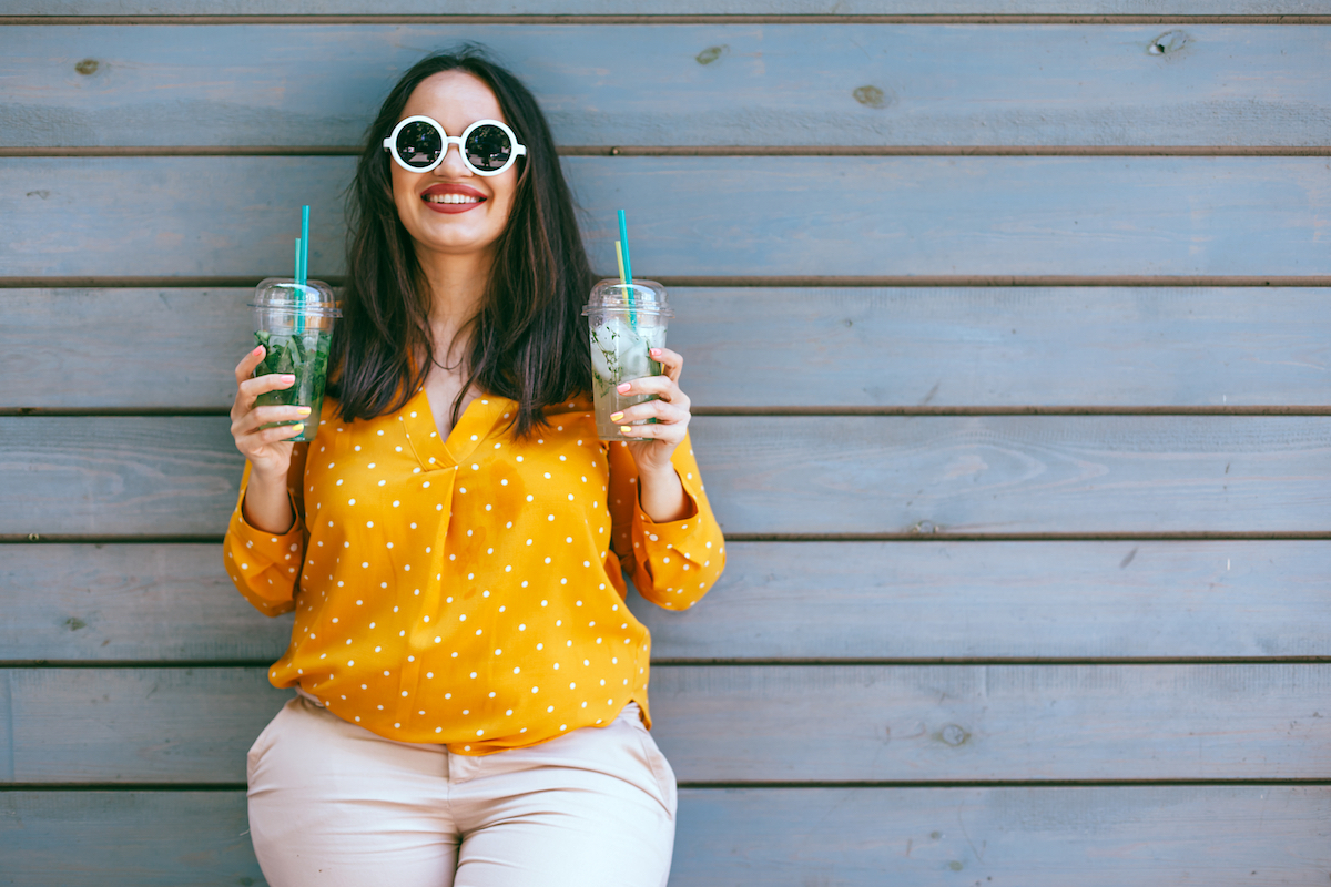 A smiling young woman in a yellow shirt standing against a wall holding two juices.