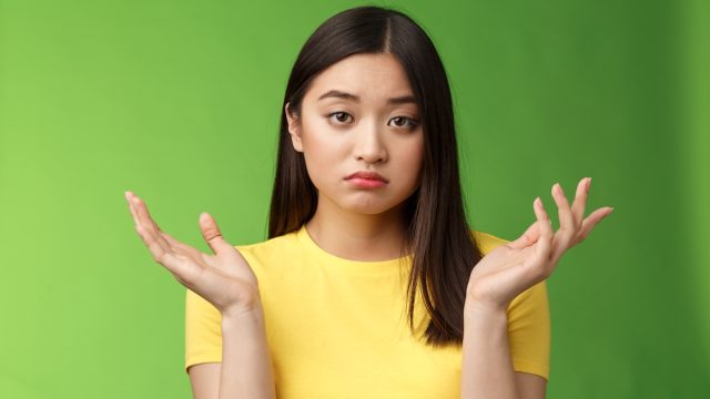Close-up of a bored-looking, unemotional young woman wearing a yellow t-shirt against a green background.
