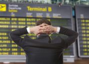A man looking at an airline departure board with his hands behind his head
