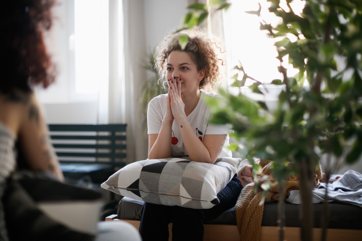 Young woman having a private conversation with her friend at home, covering mouth with surprise and listening