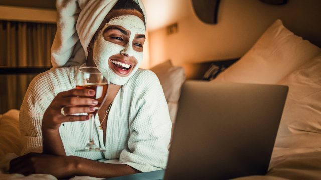 A young woman wearing a robe and a facial mask laying in bed with a glass of wine, watching something on her laptop