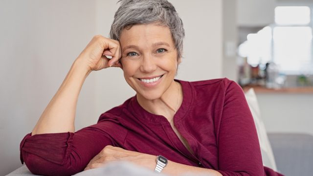 Smiling woman with short gray hair wearing a pink shirt and sitting on the couch.
