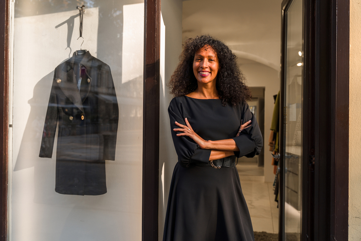 A woman standing in the entrance to a clothing store wearing a black dress and smiling with her arms crossed.