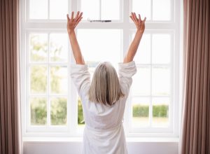 Rear view of a woman with gray hair wearing a white bathrobe stretching in the morning in front of a window