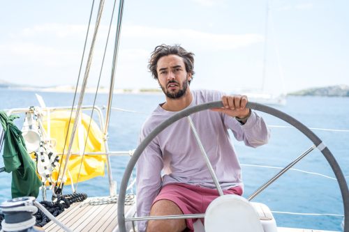 A young man wearing a light purple shirt and pink shorts steers a sailboat on the sea