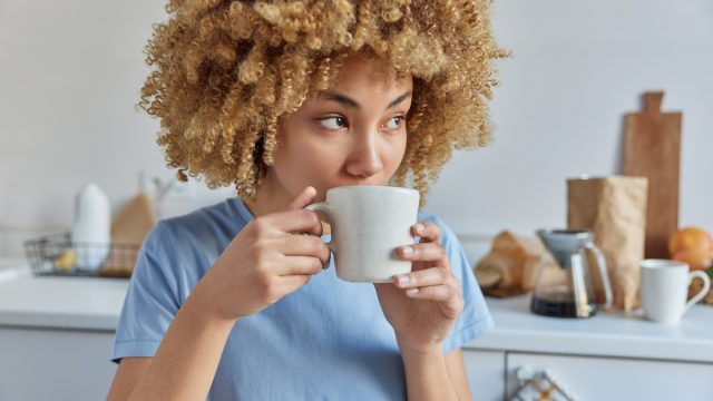 A young woman with curly hair wearing a sky-blue t-shirt drinks coffee in her kitchen