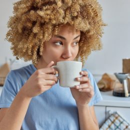 A young woman with curly hair wearing a sky-blue t-shirt drinks coffee in her kitchen