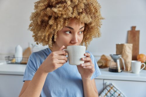 Una mujer joven con el pelo rizado y una camiseta azul cielo bebe café en su cocina