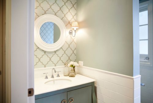 Looking into a bathroom at the vanity, which has a wallpapered wall, round white mirror, and sconce above it.