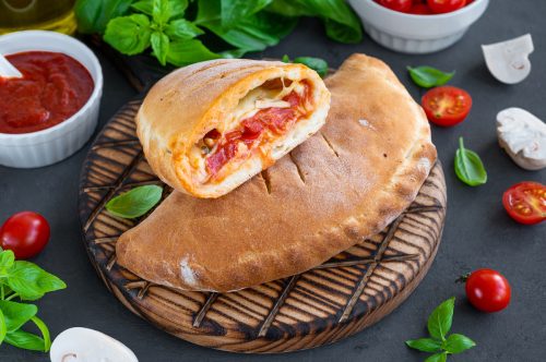 A calzone on a board on a table surrounded by fresh basil leaves, grape tomatoes, and mozzarella. 