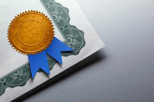 A close up of a gold seal adorned with a blue ribbon is attatched to the corner of a certificate of achievement which rests on top of a gray background which provides ample room for copy or text.