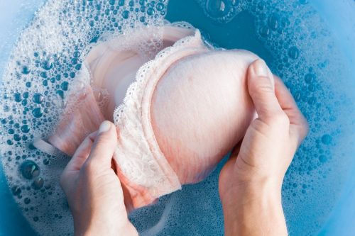 woman's hands washing light pink women's bra in a blue basin. 