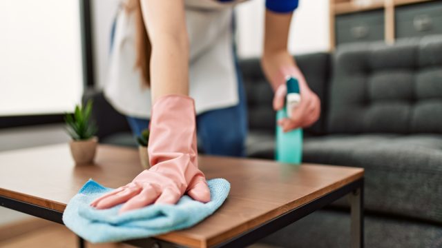 A close-up of a person wearing rubber gloves while cleaning a coffee table with a rag and cleaning product