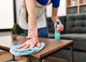 A close-up of a person wearing rubber gloves while cleaning a coffee table with a rag and cleaning product