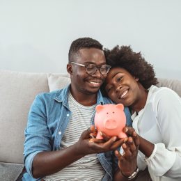 A couple sitting on the couch while smiling and holding a piggy bank