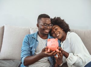 A couple sitting on the couch while smiling and holding a piggy bank