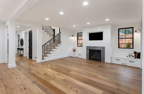 An empty farmhouse-style living room with white walls and hardwood floors, as well as a fireplace and recessed lighting