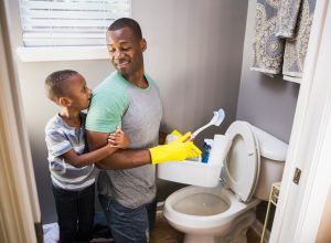 A young boy hugging the arm of a man cleaning a toilet while wearing yellow gloves and holding cleaning supplies
